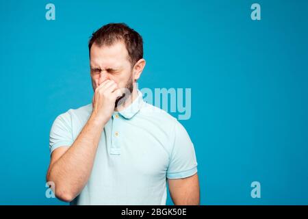 Ein Mann in einem blauen T-Shirt auf blauem Hintergrund bedeckt seine Nase mit seiner Hand. Drücken Sie die Nase gegen schlechten Geruch. Stockfoto