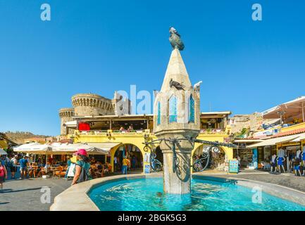Der Brunnen bei Hippokrates Platz in der Altstadt von Rhodos Griechenland mit einer Taube saß oben auf der Eule als Touristen, Shop und im Hintergrund Essen Stockfoto