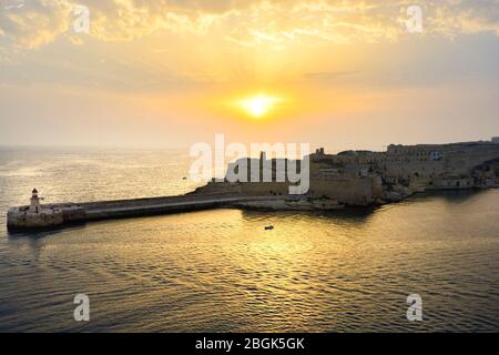 Sonnenuntergang Blick auf den Leuchtturm Ricasoli Point am Hafen von Valletta auf der Mittelmeerinsel Malta mit einem kleinen Einzelboot im Wasser. Stockfoto