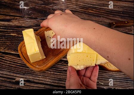 Jemand legt eine Scheibe Käse auf frischen Weizenstoast mit Butter und ein Stück Butter in eine hölzerne Butterschale auf einem hölzernen Hintergrund. Nahaufnahme Stockfoto