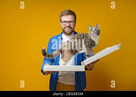 Freudiger, lächelnder Mann hält auf gelbem Hintergrund eine leere Pizzakiste in die seine liebenswerte Katze geklettert ist. Stockfoto