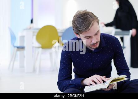Junger muslimischer Mann sitzt auf dem Boden, während er das heilige Buch Koran vor dem iftar Abendessen während eines ramadan Festes zu Hause liest Stockfoto