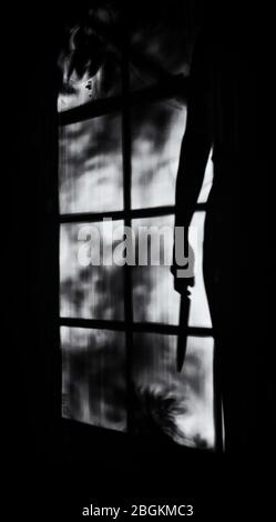 Silhouette einer Person mit einem großen Messer in der Hand, durch ein Fenster bei Nacht gesehen, Begriff von Verbrechen, Mord, Gewalt, Geheimnis Stockfoto