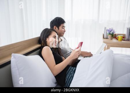 Asiatische Paar online einkaufen und mit Kreditkarte auf Laptop und Handy zu Hause bezahlen, glückliches Paar zu Hause im Netz im Bett surfen Stockfoto