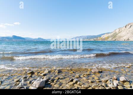 Wellen schlagen auf die felsige Küste des Okanagan Lake mit Blick auf den blauen Himmel und die ferne Bergkette Stockfoto