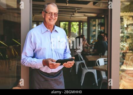 Erfolgreicher männlicher Cafe-Besitzer steht mit einem digitalen Tablet an der Tür. Älterer Mann mit Schürze arbeitet in einem Restaurant. Stockfoto