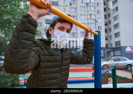 Teenager-Junge steht in der Nähe horizontale Bar von Outdoor-Trainingsgeräten, auf Spielplatz in der Nähe von Hochhäusern mit Wohnungen, eine medizinische Maske auf seinem Gesicht p Stockfoto