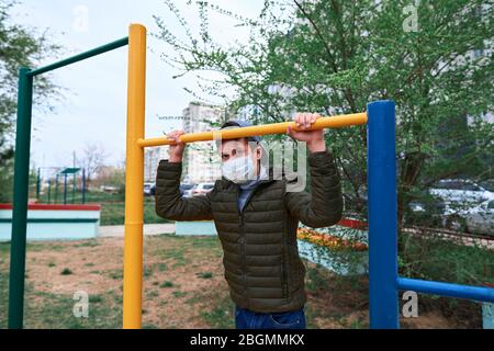 Teenager-Junge steht in der Nähe horizontale Bar von Outdoor-Trainingsgeräten, auf Spielplatz in der Nähe von Hochhäusern mit Wohnungen, eine medizinische Maske auf seinem Gesicht p Stockfoto