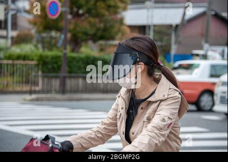 Kyoto, Japan - 12. April 2019: Junge Radfahrerin auf einer belebten Straße, die eine Anti-Umweltverschmutzung-Gesichtsmaske und ein dunkles Visier trägt, um sich vor der Sonne zu schützen. Stockfoto