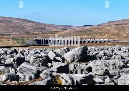 Der Zug, der Stein vom Arcow Quarry trägt, überquert das Ribblehead Viaduct, Yorkshire Dales National Park, Großbritannien. Kalksteinpflaster im Vordergrund. Stockfoto