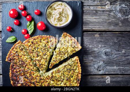 Nahaufnahme von Quiche in Scheiben mit grünen Zwiebeln und Spinat auf einem schwarzen Schiefertablett mit herzhaften Dip und Tomaten, Blick von oben, flach, leerer Raum Stockfoto