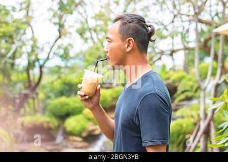 Asian Mann hält ein Glas kalten Espresso Kaffee Hintergrund verschwommene Ansichten Baum. Stockfoto