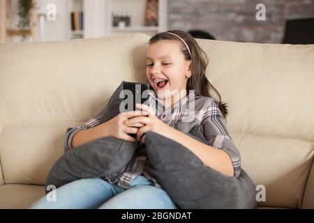 Glückliches kleines Mädchen, das auf der Couch im Wohnzimmer sitzt und ein Selfie macht. Stockfoto