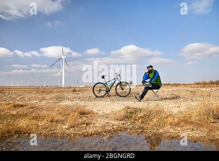 Bärtige Mann auf dem Stuhl mit seinem Mountainbike in der Nähe von Windparks in der Wüste Stockfoto