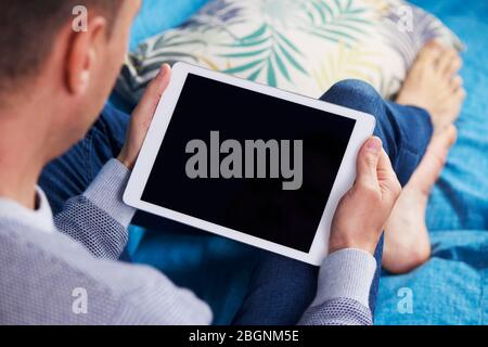 Nahaufnahme eines jungen kaukasischen Mannes, der bequem auf einer Couch liegt und seine Tafel beobachtet, mit einem schwarzen leeren Raum auf dem Bildschirm Stockfoto