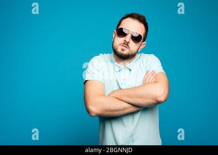 Ein Mann in Sonnenbrille ist auf dem blauen Hintergrund. Ein cooler Mann in Sonnenbrille. Ein bärtiger, cooler Kerl mit verschränkten Armen. Stockfoto