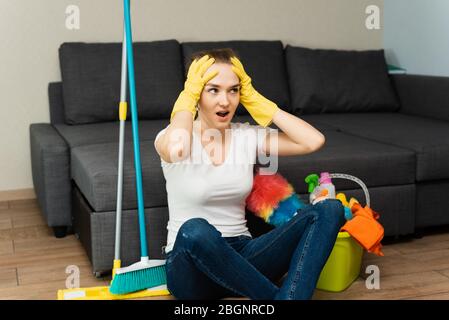 Eine schöne Frau neben Putzmitteln und Ausrüstung sitzt auf dem Boden und schreit, hält sich an ihren Kopf. Ein junges Mädchen macht die Reinigung im Haus. Stockfoto