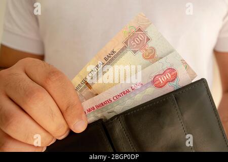 Nationale Währung der VAE. Geldscheine. Mann in weißem T-Shirt legte Dirham Banknoten in schwarzen Geldbeutel. Junger Mann hält in Händen Brieftasche mit UAE-Papier Mo Stockfoto