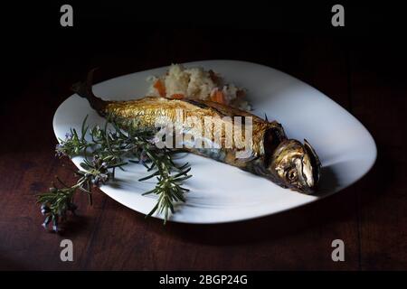 Im Ofen geröstete Makrele, serviert auf einem weißen Teller, eine kleine Portion gekochten Reis mit Karottenstücken und ein paar blühende Rosmarinzweige als Dekoration. Stockfoto