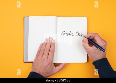 Draufsicht der Person, die Wörter SCHREIBT KAPITEL EINS auf Notizblock vor orangefarbenem Hintergrund Stockfoto