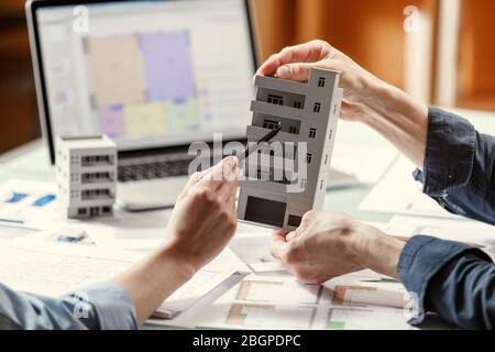 Zwei Architekten halten und diskutieren Modell eines Hauses und Architekturpläne in einem Studio mit Blaupausen und Laptop auf einem Tisch. Architektonisches Design Stockfoto