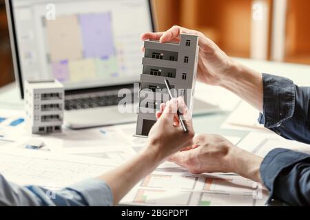 Zwei Architekten halten und diskutieren Modell eines Hauses und Architekturpläne in einem Studio mit Blaupausen und Laptop auf einem Tisch. Architektonisches Design Stockfoto