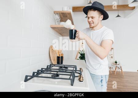 Glücklicher junger Mann gießt Kaffee aus Keramik turk in weißen Becher auf Gasherd, helle Küche Zimmer vorbereitet Stockfoto
