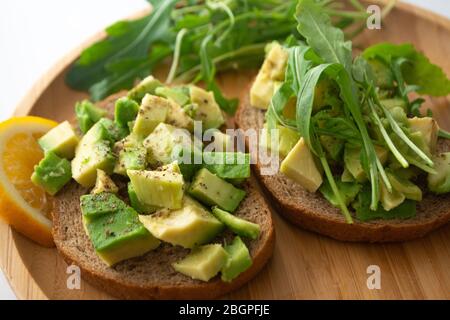 Gesundes Frühstück geröstetes Roggenbrot mit Avocado, Rucola, Zitronensaft, Salz und Pfeffer auf einem Holzteller Stockfoto