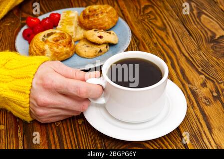 Weibliche Hände in gelben Pullover hält Tasse Kaffee und Kekse auf braunen alten Holztisch. Stockfoto