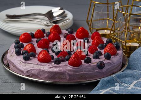 Hausgemachtes Käsekuchen mit Beeren. Käsekuchen mit Erdbeere, Heidelbeere und Himbeere auf grauem Holztisch mit Kerzen und Tellern dekoriert. Stockfoto