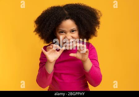Kleines schwarzes Mädchen imitiert Tiger, brüllend und grimacing über gelbem Hintergrund Stockfoto
