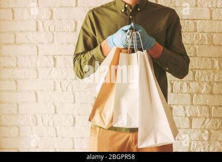 Nahaufnahme Hipster Mann trägt medizinische Schutzhandschuhe, hält Papier Einkaufstaschen in den Händen. Liefermann mit Papiertüten. Sicherer Lieferservice. Coro Stockfoto