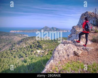 Sportliche Frau mit Rucksack auf felsigen Aussichtspunkt in Mallorka Inselberge. Rückansicht der Wandererin mittleren Alters, die auf das Meer schaut Stockfoto