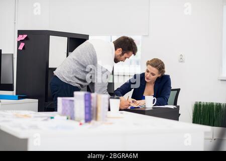 Eine Geschäftsfrau erklärt ihrem Mitarbeiter etwas, während sie an ihrem Schreibtisch sitzt. Der Mitarbeiter hört aufmerksam zu und lehnt sich nach vorne. Stockfoto