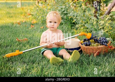 Portrait blonde Kind Junge spielt mit Sprüher und Trauben im Garten. Glückliche Kindheit, Sommerferien im Dorf. Schöne kaukasische Kind Junge Bauer 2 Stockfoto