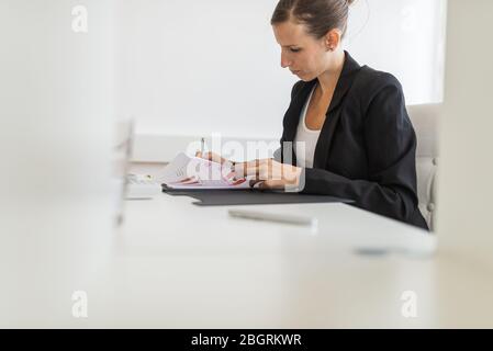 Junge Geschäftsfrau sitzt an ihrem hellen Büroschreibtisch, arbeitet an Papierkram und Finanzbericht in Diagrammen und Diagrammen dargestellt. Stockfoto