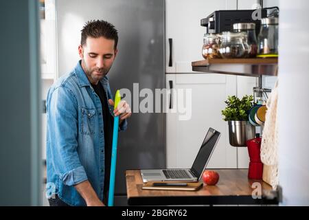 Moderner Mann mit Besen fegenden Boden in seiner Küche zu Hause. Mann, der Aufgaben übernimmt, die normalerweise von Frauen erledigt werden. Reinigung, Hausarbeit und Housekeeping Konzept Stockfoto