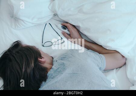 Kranker junger Mann schläft im Bett mit Brille und Thermometer neben ihm auf dem Bett Stockfoto
