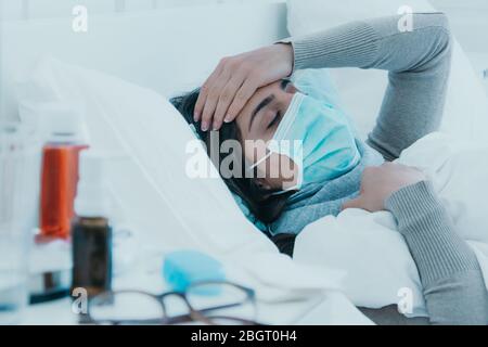 Junge kranke Frau liegt müde im Bett mit einer Gesichtsmaske und hält ihren Kopf wegen Kopfschmerzen. Stockfoto