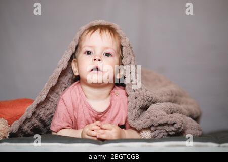 Ein einjähriger Junge versteckt sich unter einer beigefarbenen Strickdecke oder einem Bettüberwurf. Ein Junge im Bett. Stockfoto