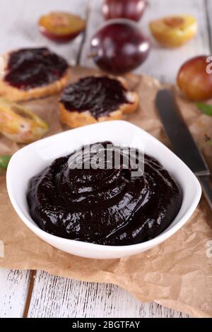 Pflaumenmarmelade, Scheiben Brot mit Pflaumenmarmelade und frischen Pflaumen in Glasform auf einem Stück Papier auf Holztisch auf hellem Hintergrund Stockfoto