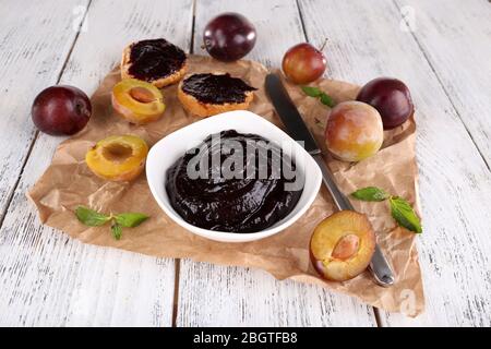 Pflaumenmarmelade, Scheiben Brot mit Pflaumenmarmelade und frischen Pflaumen in Glasform auf einem Stück Papier auf Holztisch auf hellem Hintergrund Stockfoto