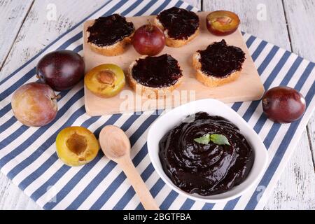 Schüssel mit Pflaumenmarmelade, Scheiben Brot mit Pflaumenmarmelade und frischen Pflaumen auf Schneidebrett auf Serviette auf Holzhintergrund Stockfoto