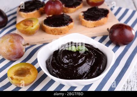 Schüssel mit Pflaumenmarmelade, Scheiben Brot mit Pflaumenmarmelade und frischen Pflaumen auf Schneidebrett auf Serviette auf Holzhintergrund Stockfoto