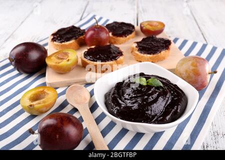 Schüssel mit Pflaumenmarmelade, Scheiben Brot mit Pflaumenmarmelade und frischen Pflaumen auf Schneidebrett auf Serviette auf Holzhintergrund Stockfoto