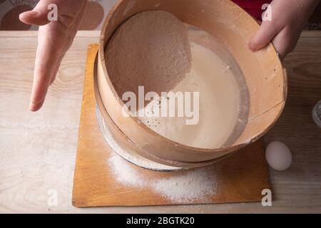 Große Schüssel, Schneidebrett, Sieb zum Sieben von Mehl auf dem Tisch. Weibliche Hände sieben Mehl in der Küche. Stockfoto