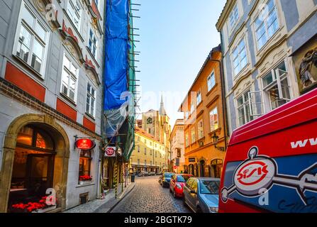 Eine enge Straße mit Geschäften, gesäumt von Autos und Lastwagen in Prag, Tschechische Republik, während Sonnenlicht einen Kirchturm im Stadtteil Male Strana hervorhebt Stockfoto