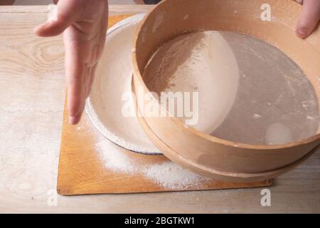 Große Schüssel, Schneidebrett, Sieb zum Sieben von Mehl auf dem Tisch. Weibliche Hände sieben Mehl in der Küche. Stockfoto