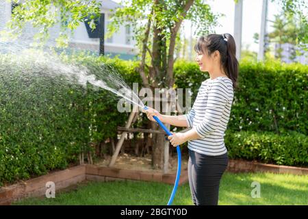 Asiatische Frau glücklich in Haus Hinterhof Sommergarten, mit Gartenschlauch Spritzwasser auf dem Rasen und Baum Blätter während des Aufenthalts zu Hause mit kostenlos Stockfoto