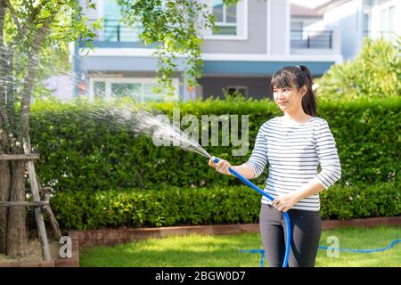 Asiatische Frau glücklich in Haus Hinterhof Sommergarten, mit Gartenschlauch Spritzwasser auf dem Rasen und Baum Blätter während des Aufenthalts zu Hause mit kostenlos Stockfoto